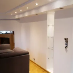 Jasne wnętrze salonu z drewnianą podłogą, kominkiem i szklanymi półkami w ścianie, widoczne gniazdka elektryczne przed montażem osprzętu.