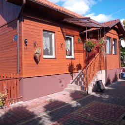 Grzegorz Szynalik - Drewniany dom z pomalowaną elewacją w odcieniach brązu, z widocznymi numerem domu, kwiatami w wiszących doniczkach i schodami wejściowymi.