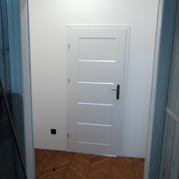 Grzegorz Szynalik - Białe drzwi z poziomymi panelami w białej ścianie, widoczne zza niebieskiej framugi. Parkiet w jodełkę na podłodze. Minimalistyczny design.