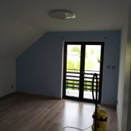 Pokój po malowaniu ścian na niebiesko, z drewnopodobną podłogą i balkonem. Żółty odkurzacz stoi przy ścianie. Widoczne gniazdka elektryczne i lampa sufitowa.