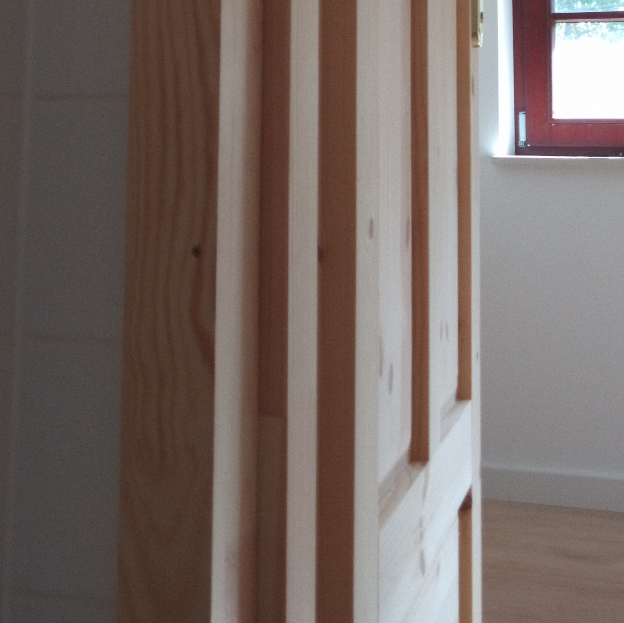 Fragment drewnianej konstrukcji, prawdopodobnie drzwi lub ścianki działowej, z widocznymi pionowymi listwami i oknem w tle.