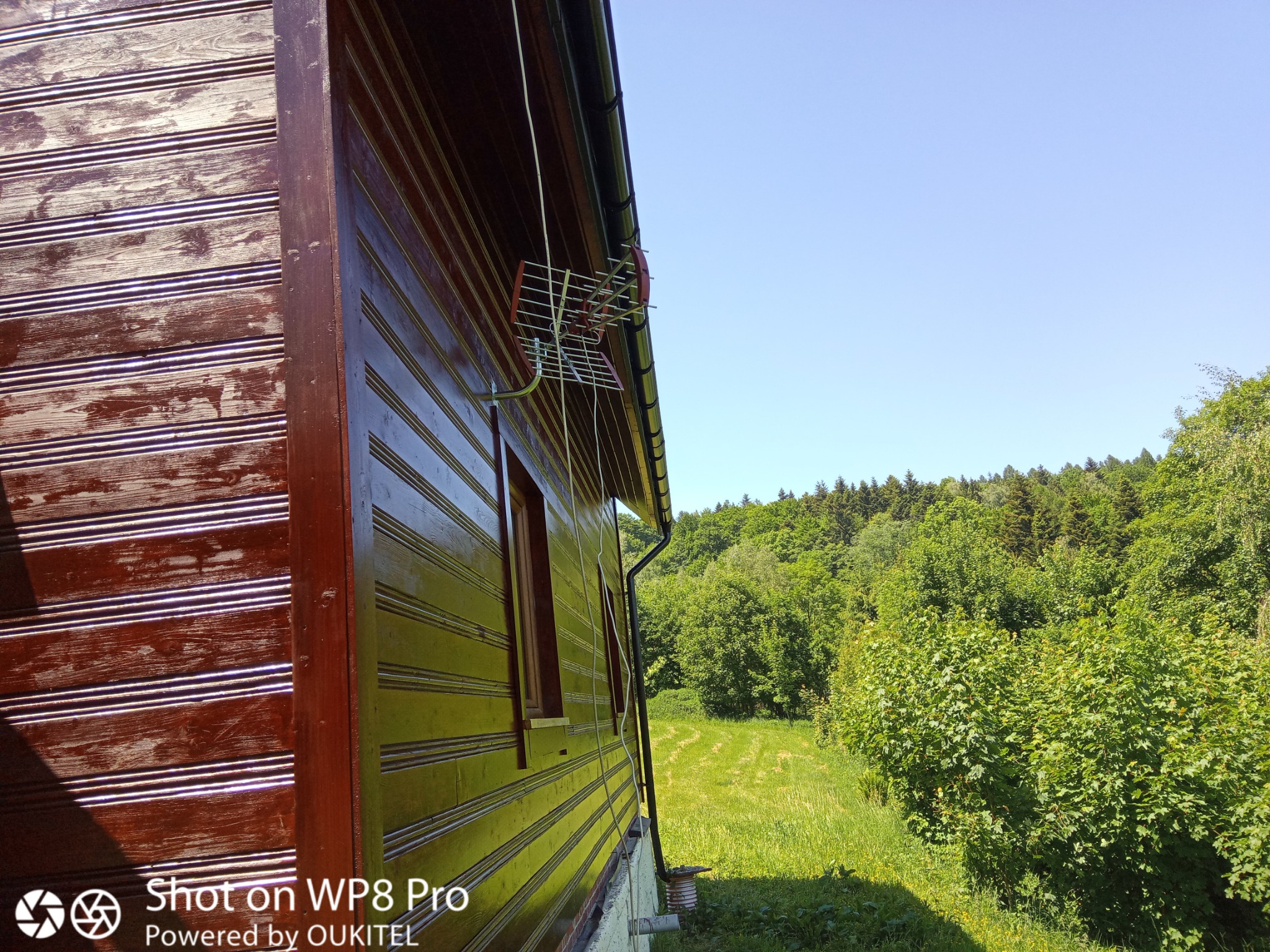 Boczny widok drewnianego domu z brązowymi deskami, widoczna antena, rynna i okno, w tle zielony krajobraz z drzewami i niebem.
