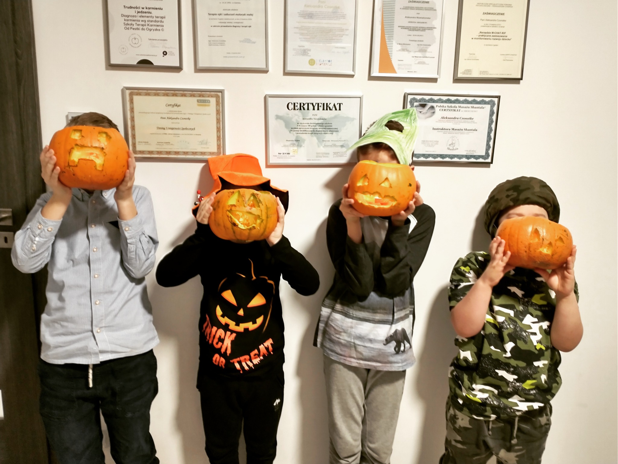 Czterech chłopców trzyma przed twarzami wydrążone dynie z wyciętymi wzorami na tle ściany z certyfikatami i dyplomami.