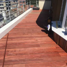 Wykończony taras na dachu budynku z deskami w odcieniach czerwieni, widoczna balustrada i panorama miasta w tle.