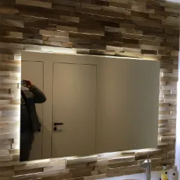 Łazienka z lustrem podświetlanym LED otoczonym dekoracyjną ścianą z drewnianych paneli, widoczne elementy armatury i odbicie osoby robiącej zdjęcie.