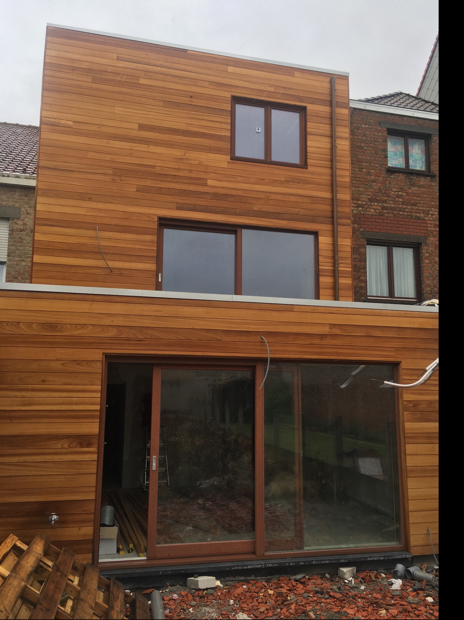 Nowoczesna elewacja budynku obłożona panelami z drewna egzotycznego, z widocznymi oknami z brązowymi ramami i elementami wykończeniowymi w kolorze srebrnym, w tle fragment sąsiedniego budynku...