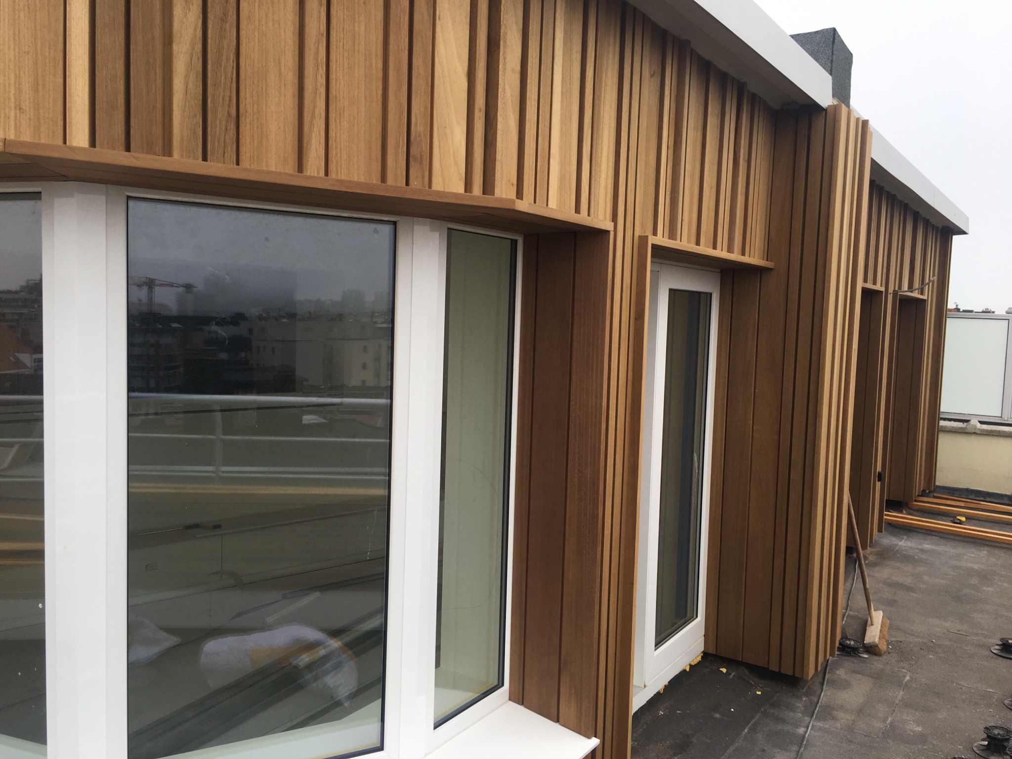 Elewacja budynku wykończona pionowymi panelami z drewna, z białymi ramami okiennymi i drzwiowymi, widok z zewnątrz na dachu, z widoczną panoramą miasta w odbiciu okna.