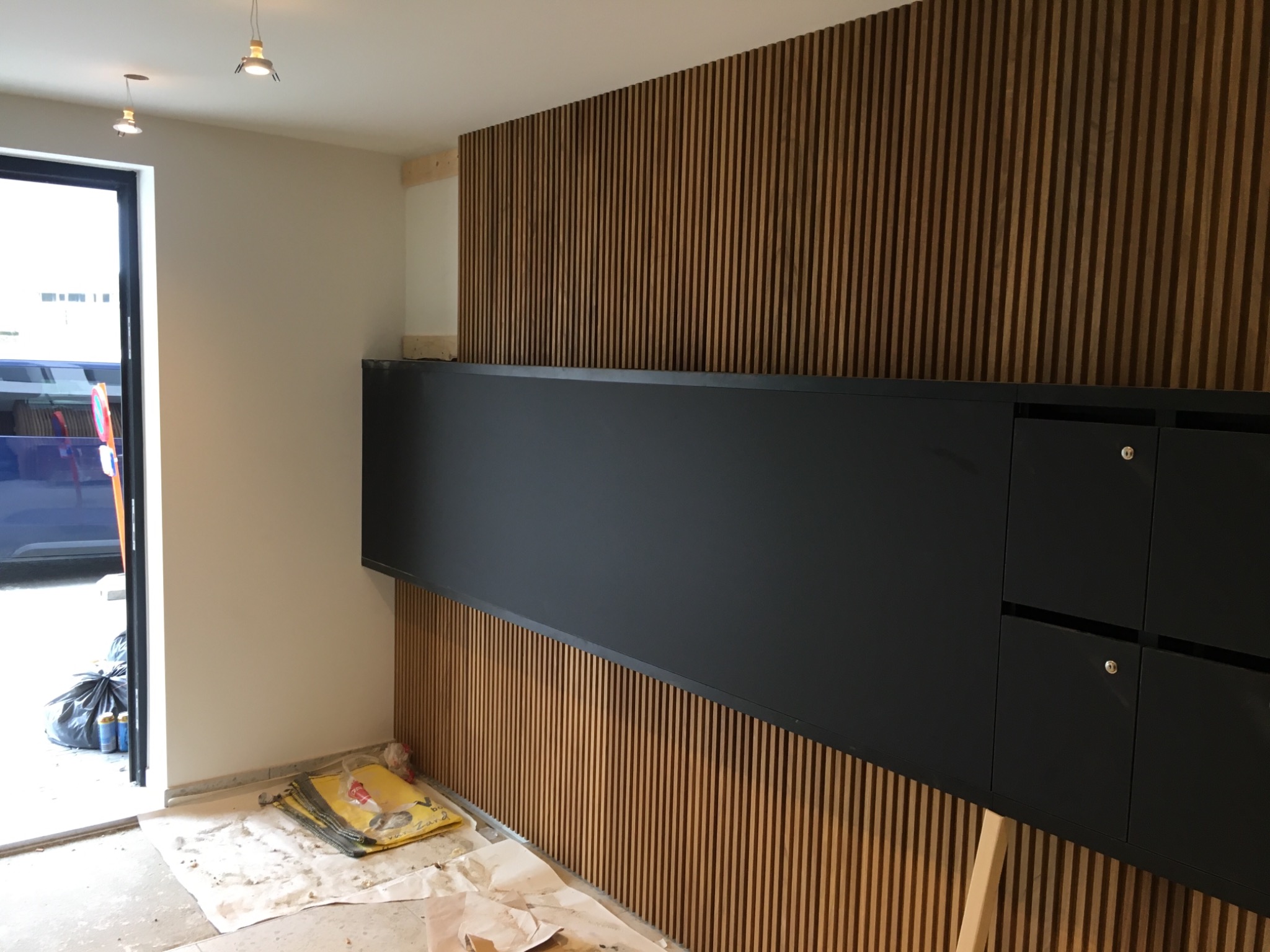 Montaż drewnianych paneli ściennych z wbudowaną czarną szafką, widoczne narzędzia i zabezpieczenia na podłodze, otwarte drzwi wejściowe z widokiem na zewnątrz.