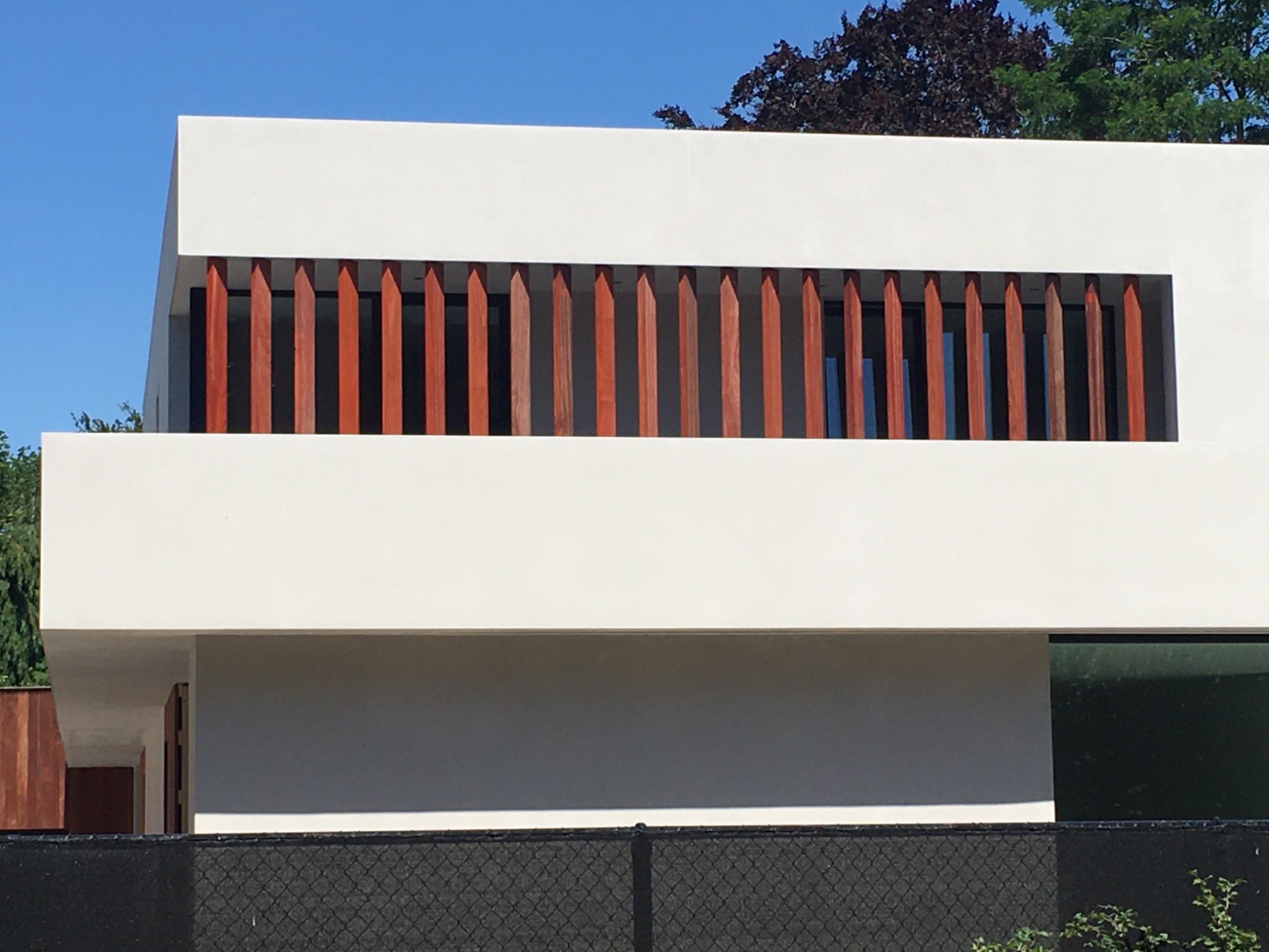 Nowoczesny, biały budynek z drewnianymi, pionowymi przesłonami na balkonie, widoczny fragment czarnego ogrodzenia i zieleń drzew w tle.
