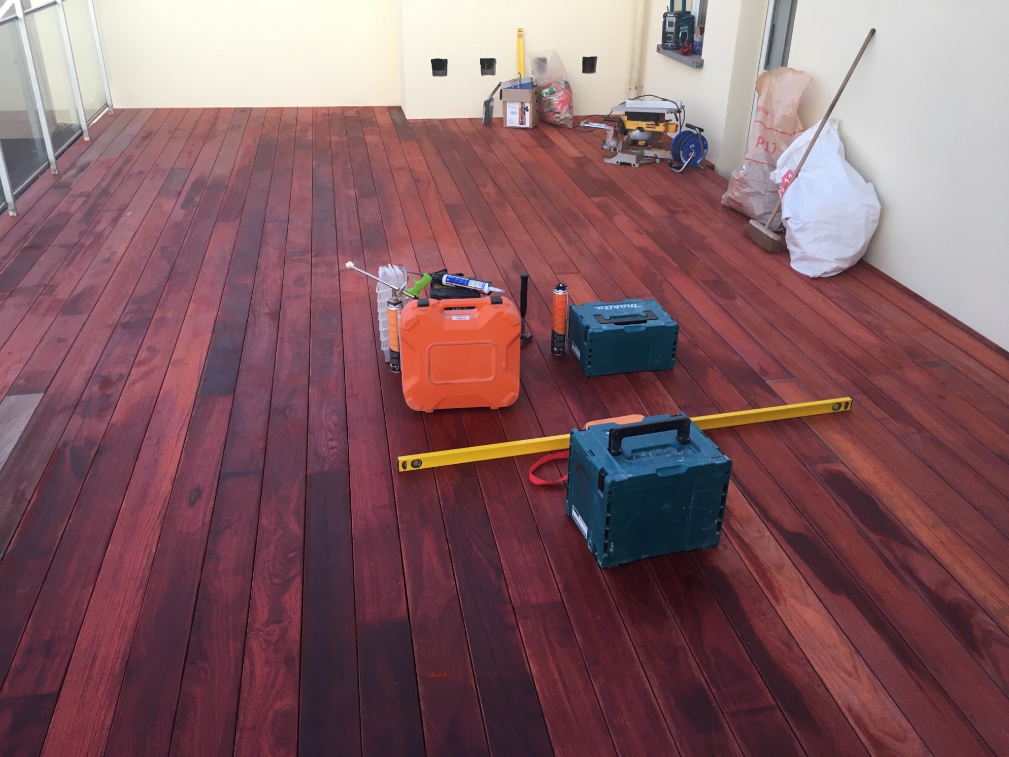 Ciemnoczerwony taras kompozytowy w trakcie montażu, widoczne narzędzia: poziomica, skrzynki narzędziowe Makita, pistolet do silikonu i torby z materiałami budowlanymi w tle.