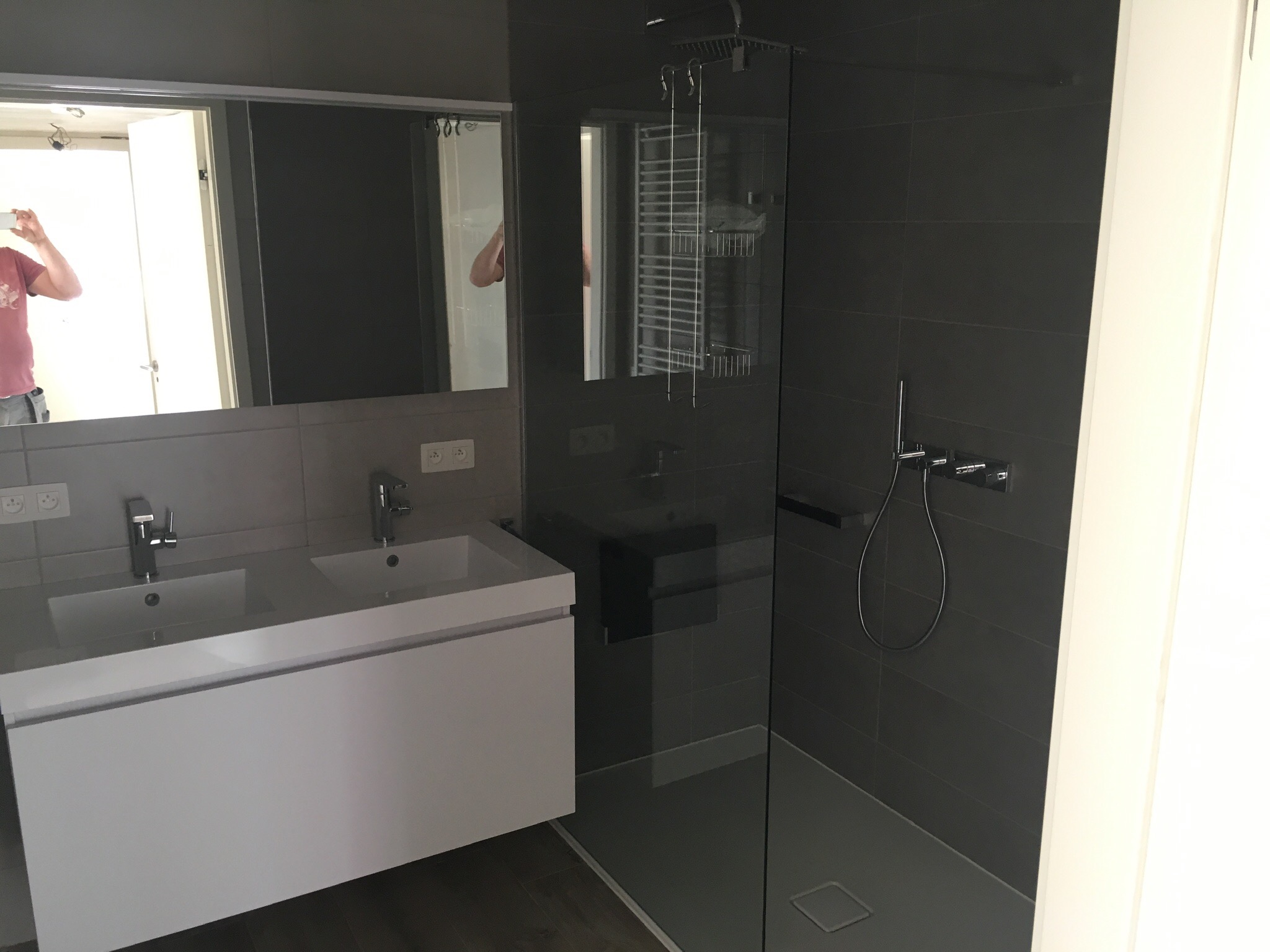 Nowoczesna łazienka z szarymi płytkami, kabiną prysznicową typu walk-in z deszczownicą i białą szafką z dwiema umywalkami, widok z boku, w lustrze odbija się osoba robiąca zdjęcie.