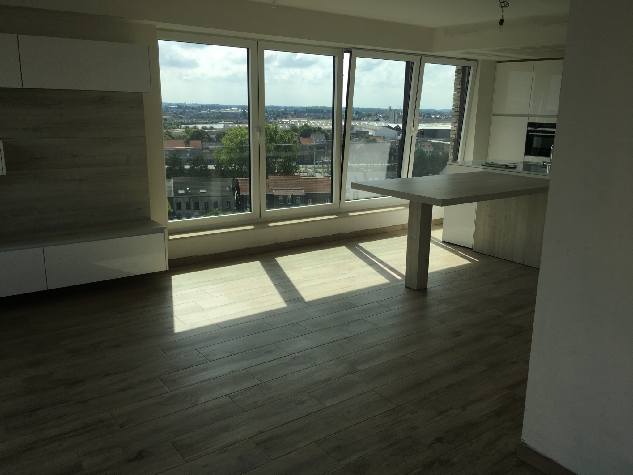 Jasne wnętrze mieszkania z dużymi oknami wychodzącymi na miasto, drewnopodobna podłoga, białe meble kuchenne i wysoki stół z blatem w kolorze szarym.