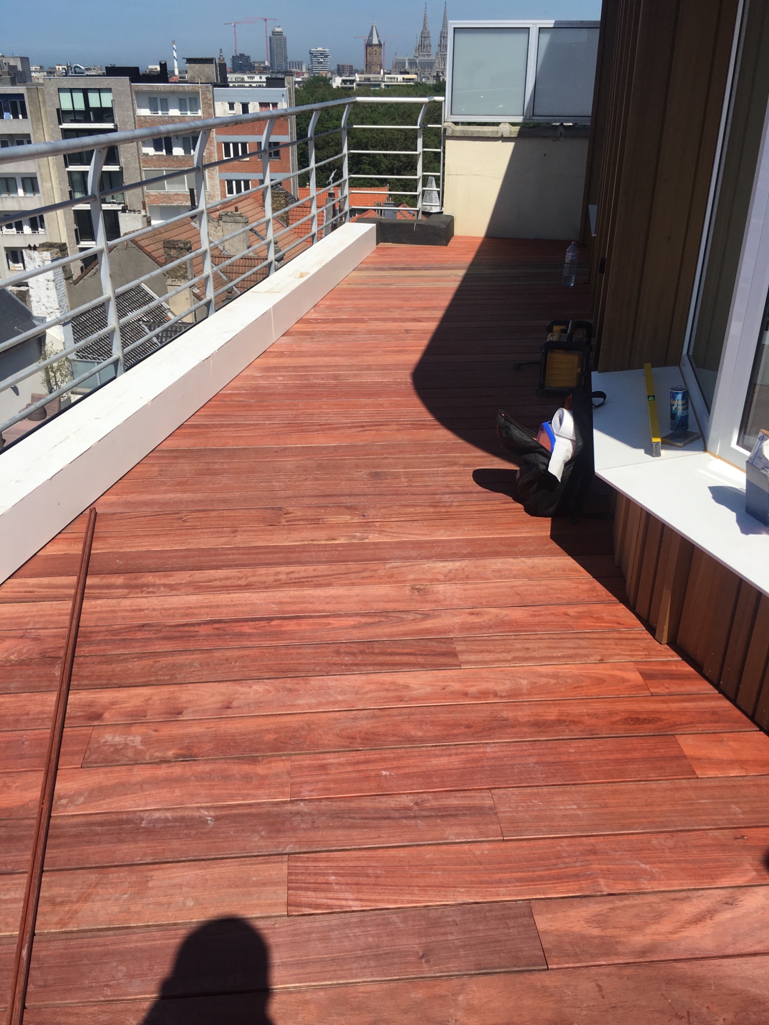 Wykończony taras na dachu budynku z deskami w odcieniach czerwieni, widoczna balustrada i panorama miasta w tle.