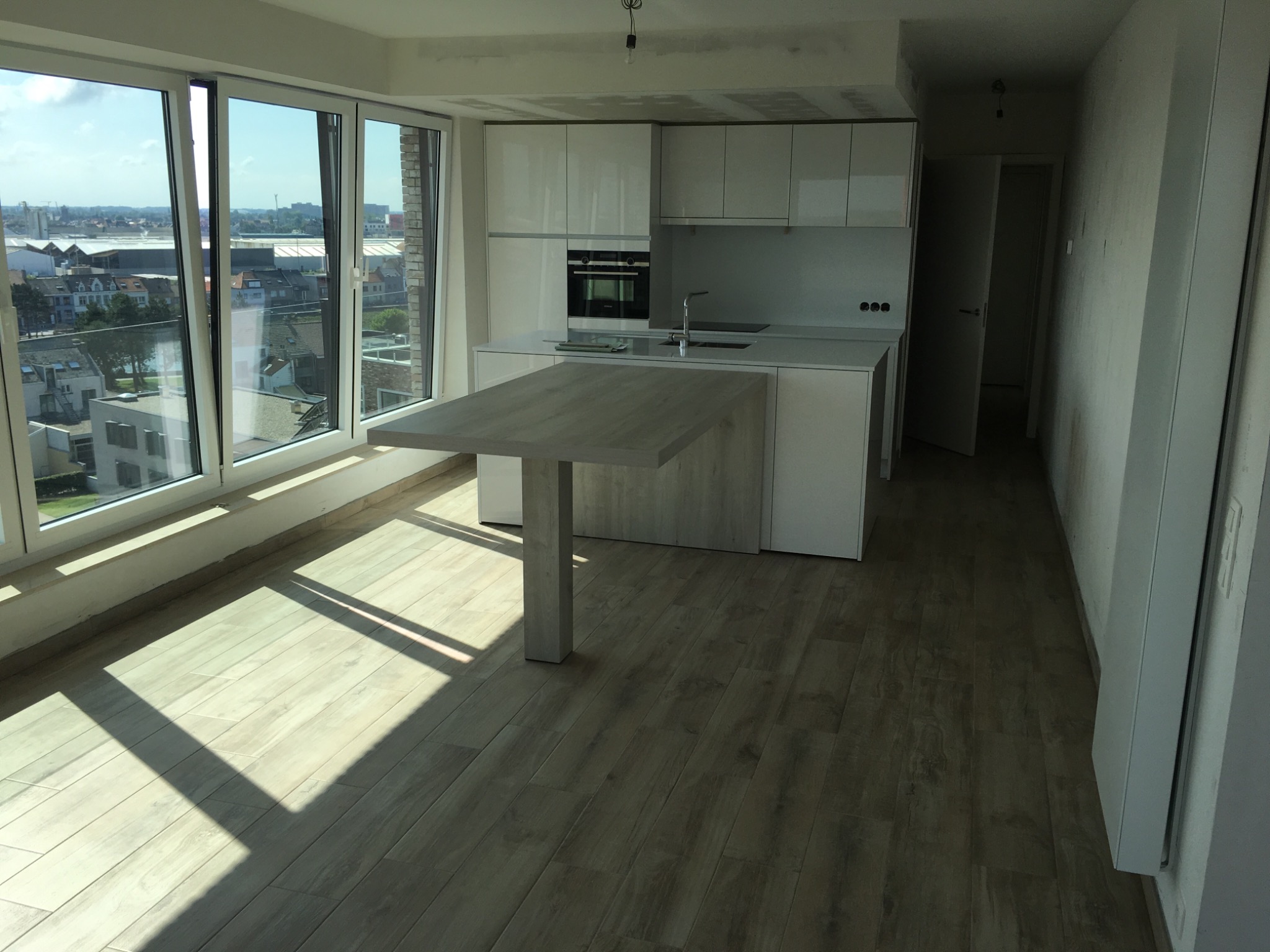 Wnętrze jasnego, nowoczesnego mieszkania w stanie deweloperskim z widokiem na miasto przez duże okna, widoczna kuchnia z wyspą i drewnopodobna podłoga w promieniach słońca.