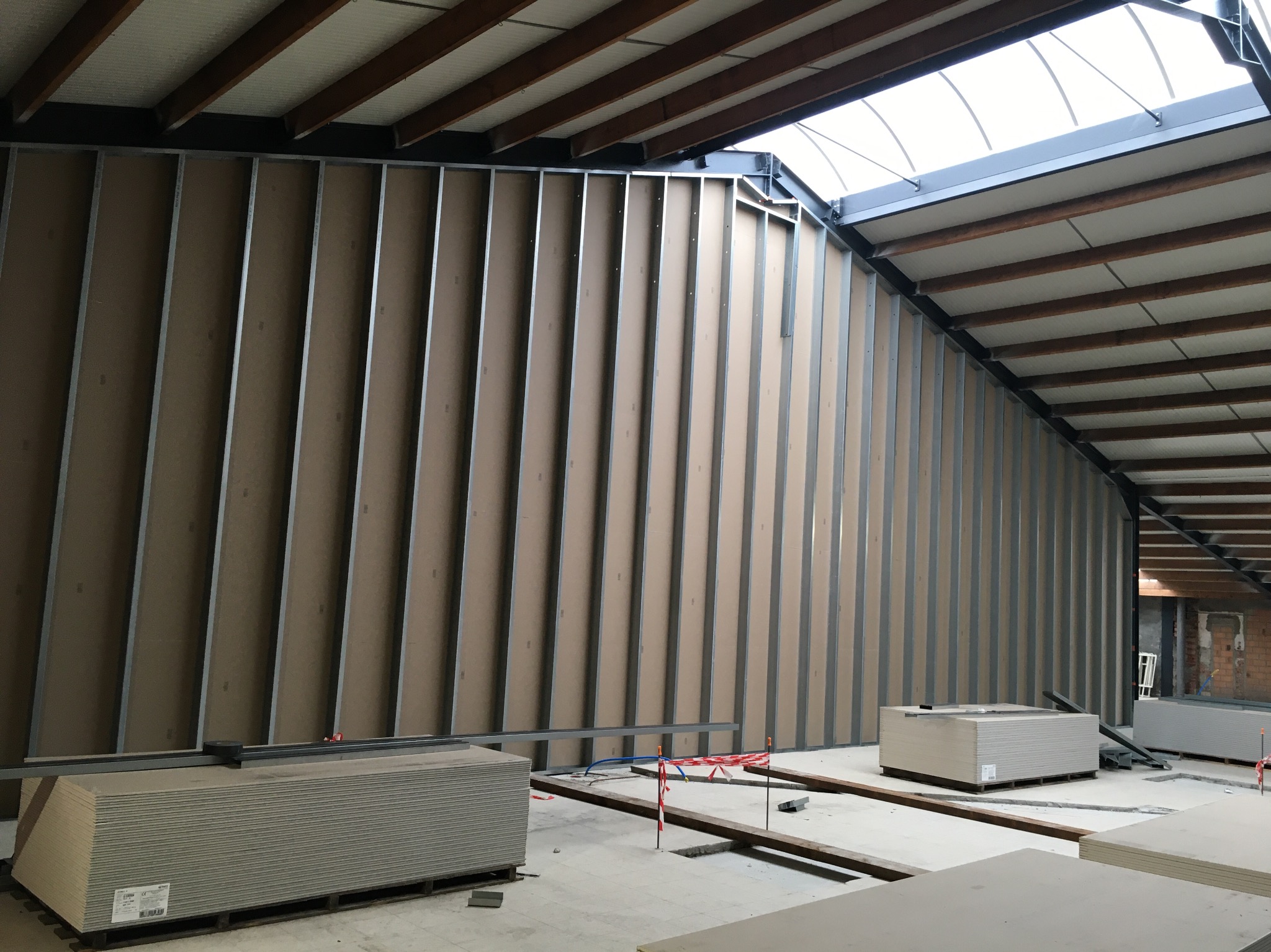 Montaż metalowej konstrukcji ścian działowych z płyt kartonowo-gipsowych w wysokim pomieszczeniu z oknem dachowym, widoczne palety z płytami gipsowymi.