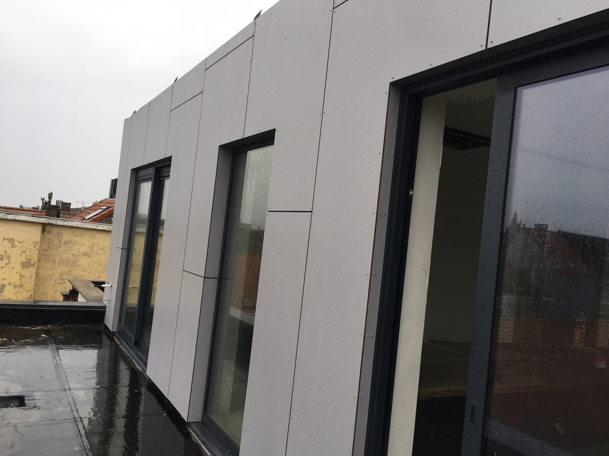 Szara elewacja budynku z panelami, widoczne okna z czarnymi ramami i mokry, czarny dach płaski po deszczu.