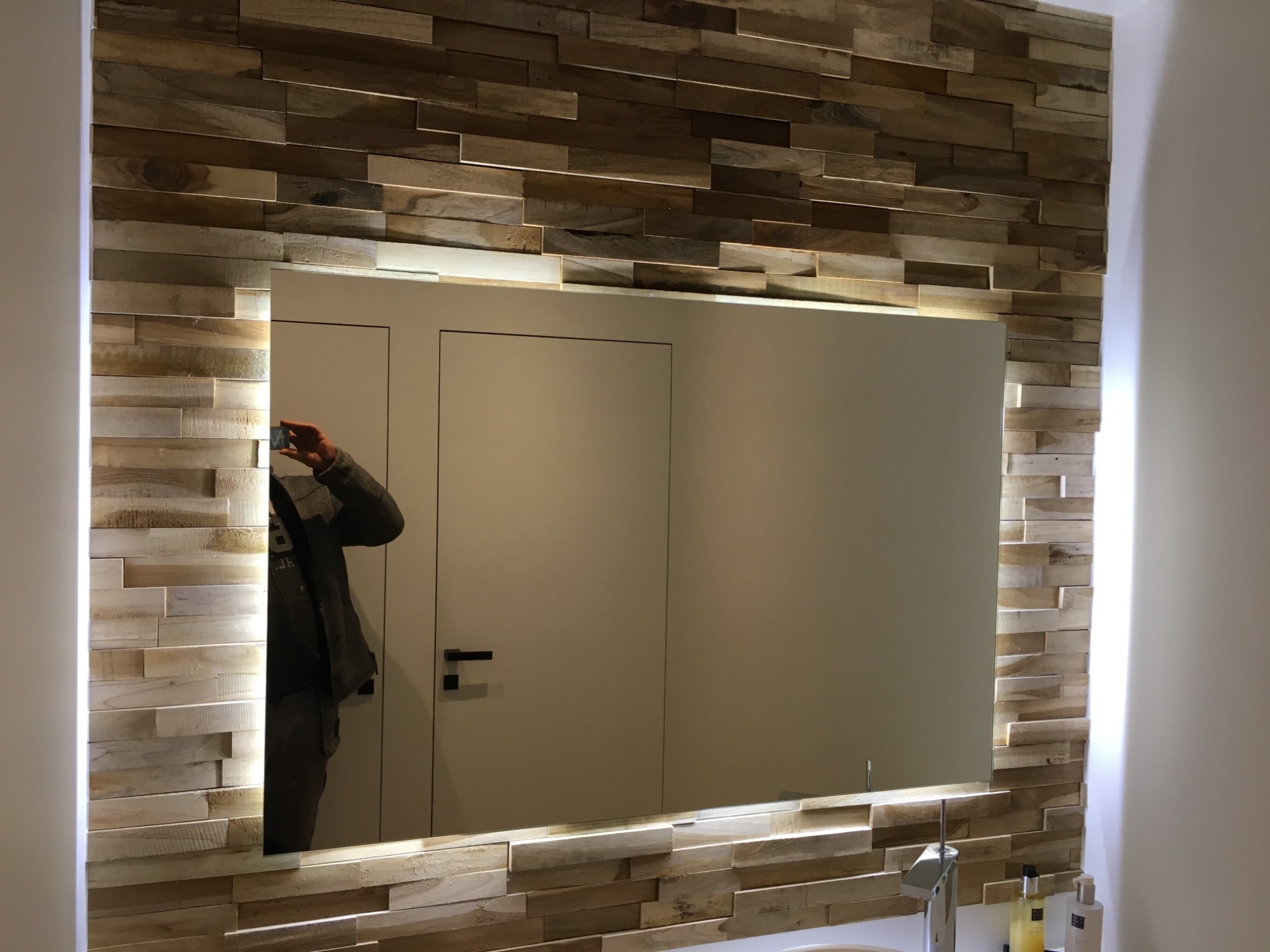 Łazienka z lustrem podświetlanym LED otoczonym dekoracyjną ścianą z drewnianych paneli, widoczne elementy armatury i odbicie osoby robiącej zdjęcie.