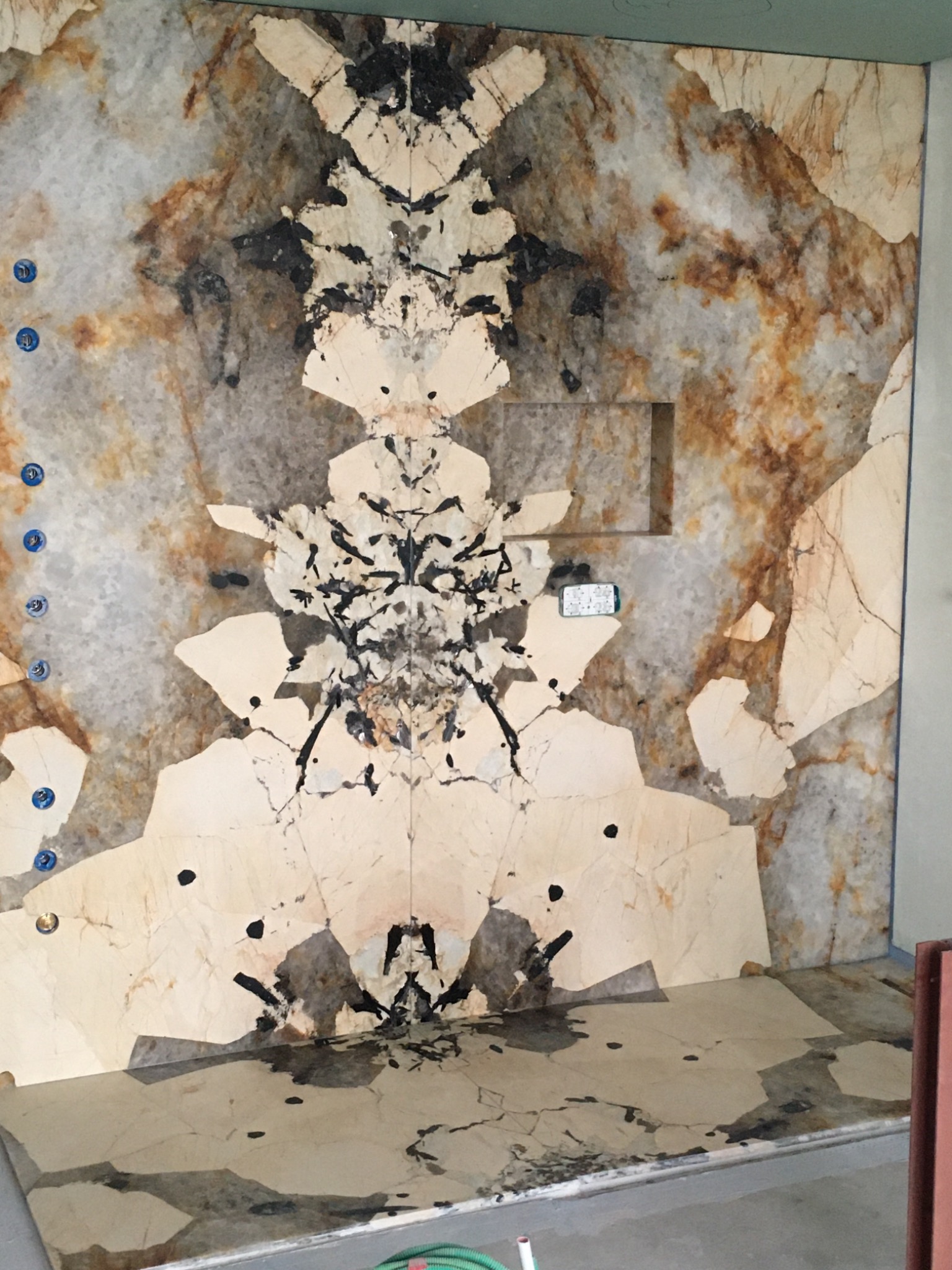 Ściana i podłoga łazienki wyłożone płytami z wzorzystego kamienia naturalnego, widoczne otwory na armaturę, wnęka w ścianie i ślady po kleju lub zaprawie na podłodze.