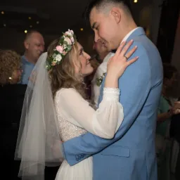 Para młoda w objęciach podczas pierwszego tańca. Panna młoda w wianku z kwiatów i welonie, pan młody w błękitnym garniturze. Uśmiechnięci, patrzą na siebie.