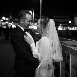 Mateusz Alfut - Czarno-białe zdjęcie uśmiechniętej pary młodej na molo nocą, z widocznymi światłami w tle. Panna młoda w welonie, pan młody w smokingu z muchą.