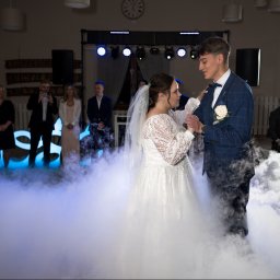 Mateusz Alfut - Para młoda w tańcu, otoczona dymem na parkiecie. W tle goście robiący zdjęcia, dekoracje świetlne i zegar na ścianie. Ujęcie z reportażu ślubnego.