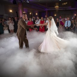 Mateusz Alfut - Para młoda w tańcu na parkiecie wypełnionym dymem, otoczona gośćmi weselnymi. Panna młoda w sukni z aplikacjami, pan młody w brązowym garniturze. Dekoracje sali.