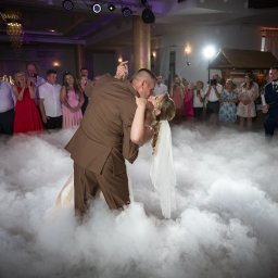 Mateusz Alfut - Pierwszy taniec pary młodej w objęciach, otulonych sztuczną mgłą, na tle gości weselnych z telefonami i dekoracji sali.