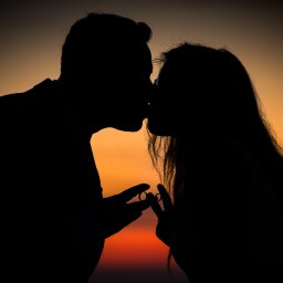 Mateusz Alfut - Silueta całującej się pary na tle zachodzącego słońca. W dłoniach trzymają obrączki ślubne, tworząc romantyczny i intymny moment.