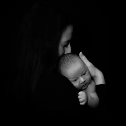 Matka w czarnym ubraniu delikatnie całuje noworodka w czarno-białej tonacji, dziecko patrzy w dół z zaciśniętą piąstką.
