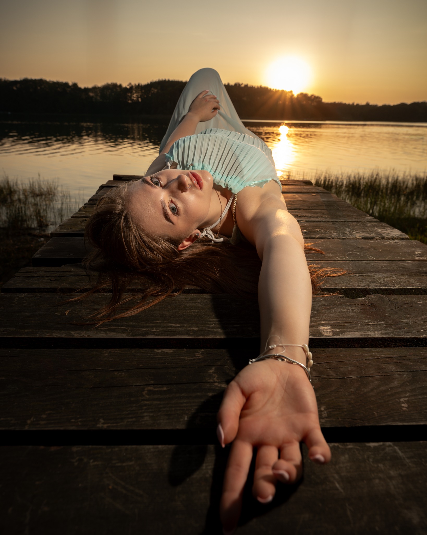 Młoda kobieta w zwiewnej, błękitnej sukience leży na drewnianym molo nad jeziorem, z zachodzącym słońcem w tle, eksponując biżuterię na szyi i nadgarstku.