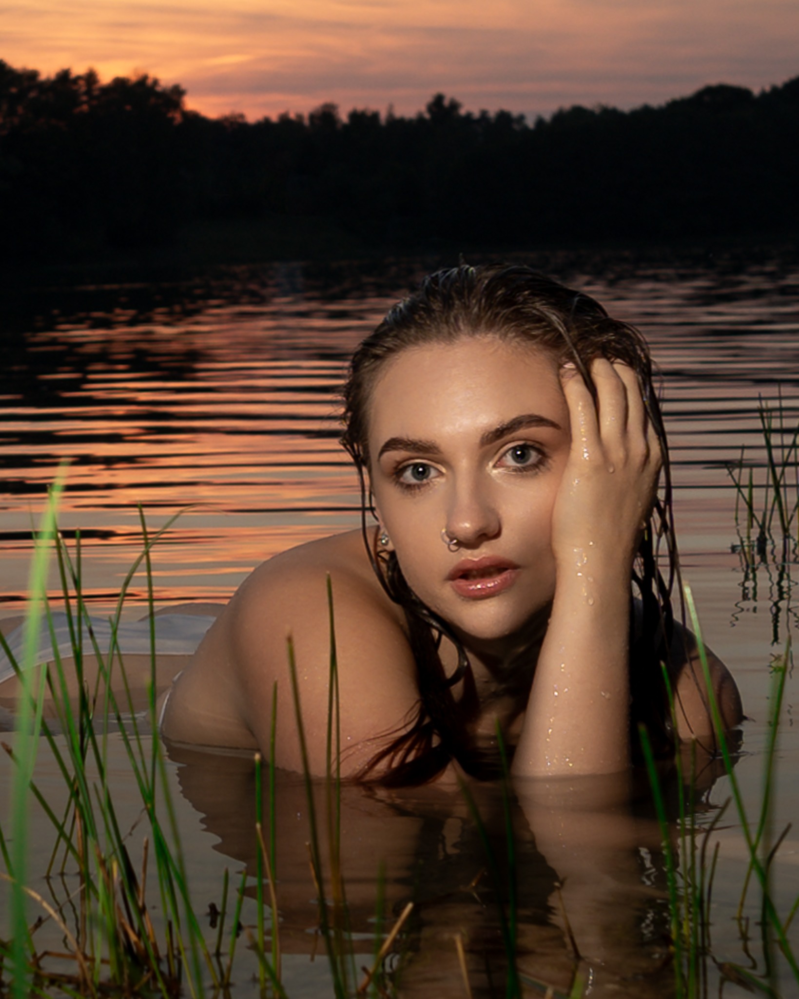 Portret kobiety zanurzonej w wodzie po ramiona, z mokrymi włosami opadającymi na twarz i ramiona, dłoń przy twarzy, trzciny w pierwszym planie, w tle zachodzące słońce odbijające się w tafli wody.