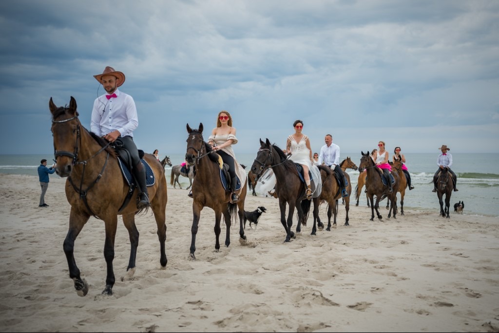 Panna młoda i pan młody na koniach podczas sesji ślubnej na plaży. Nietypowe ujęcie z konnej sesji plenerowej. W tle widać morze i pozostałych gości.