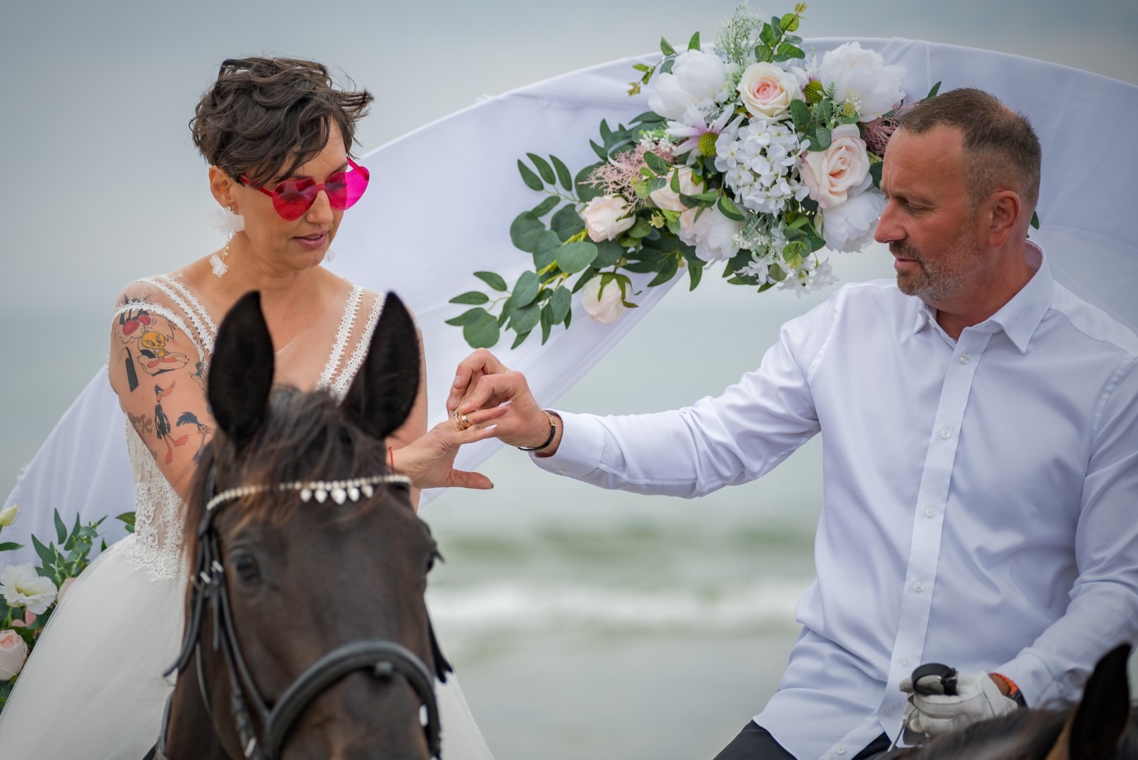 Para wymienia obrączki podczas ślubu na koniach, na tle plaży i dekorowanego łuku. Panna młoda w sukni, z tatuażem i okularach w kształcie serc.