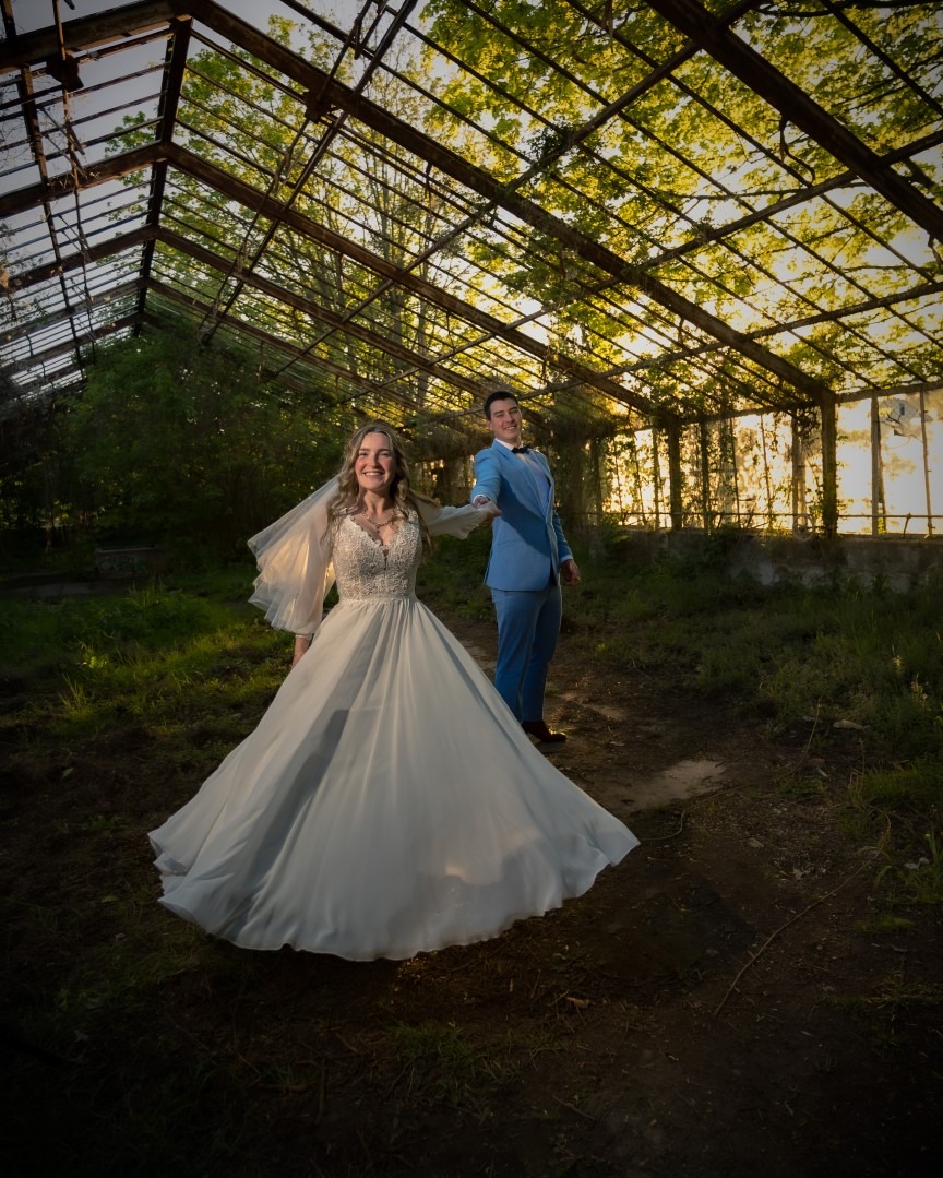 Para młoda w ruinach szklarni: uśmiechnięta panna młoda w koronkowej sukni, pan młody w błękitnym garniturze, zielona roślinność w tle, ciepłe światło zachodzącego słońca.