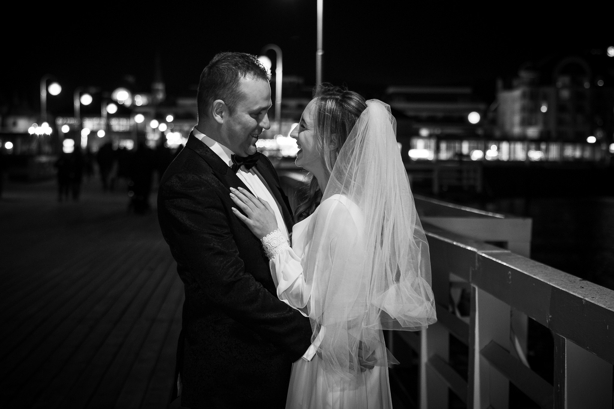 Czarno-białe zdjęcie uśmiechniętej pary młodej na molo nocą, z widocznymi światłami w tle. Panna młoda w welonie, pan młody w smokingu z muchą.