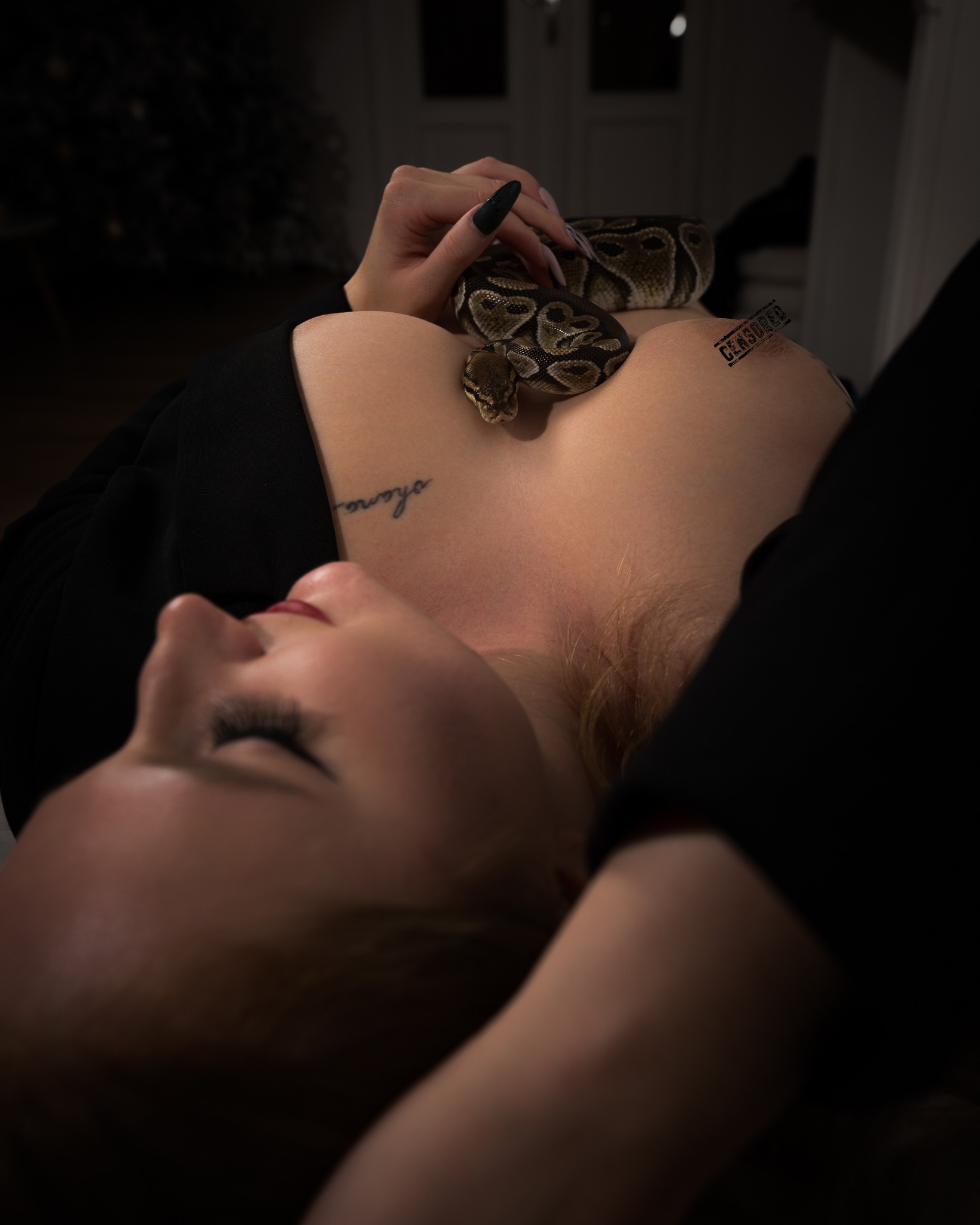 Kobieta leżąca na plecach z wężem boa na dekolcie, widoczny tatuaż na ramieniu, ciemne tło, oświetlenie studyjne, długie paznokcie.