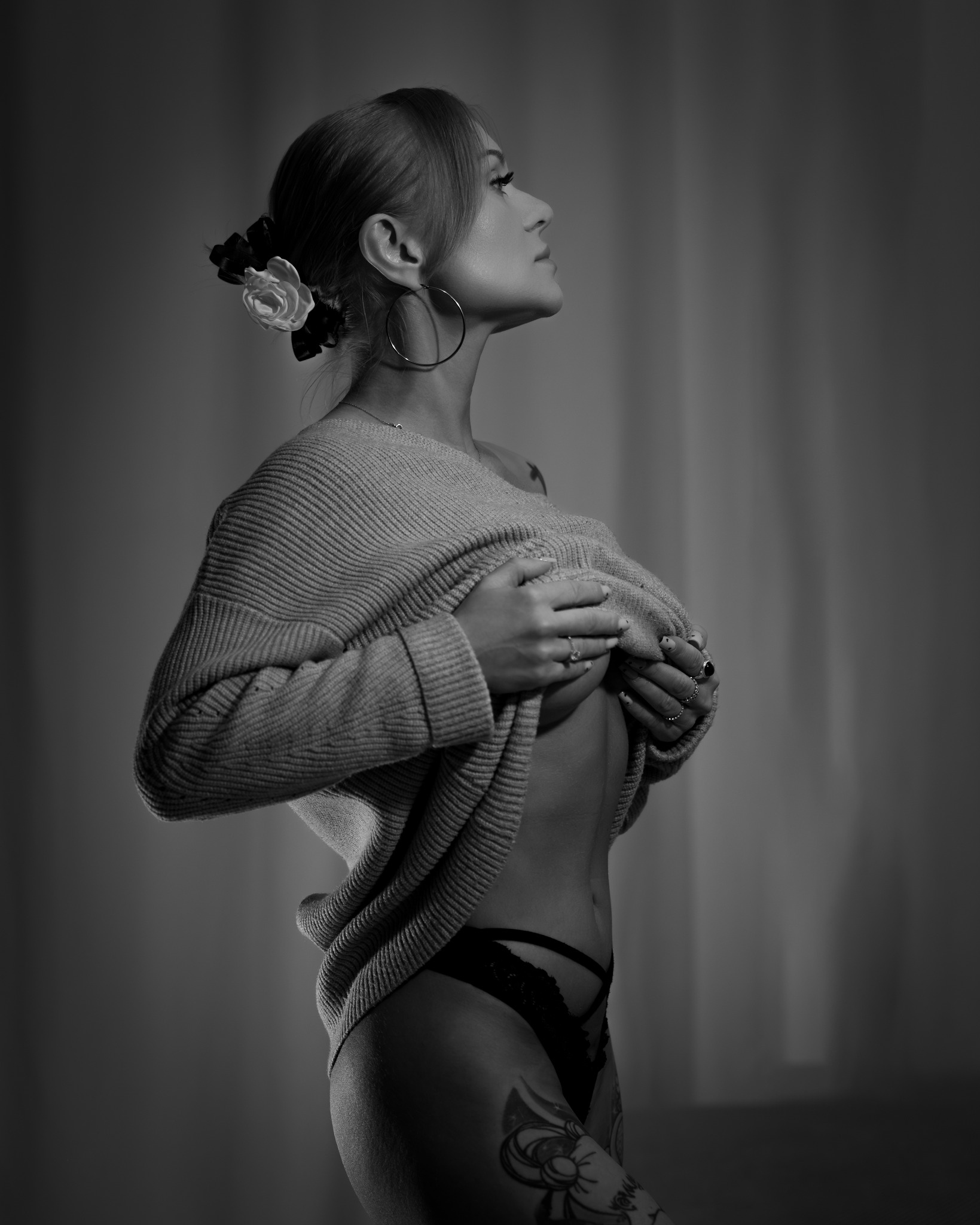 Czarno-biały portret kobiety z kwiatem we włosach, w swetrze, z widocznym tatuażem na udzie, spoglądającej w górę, na neutralnym tle.