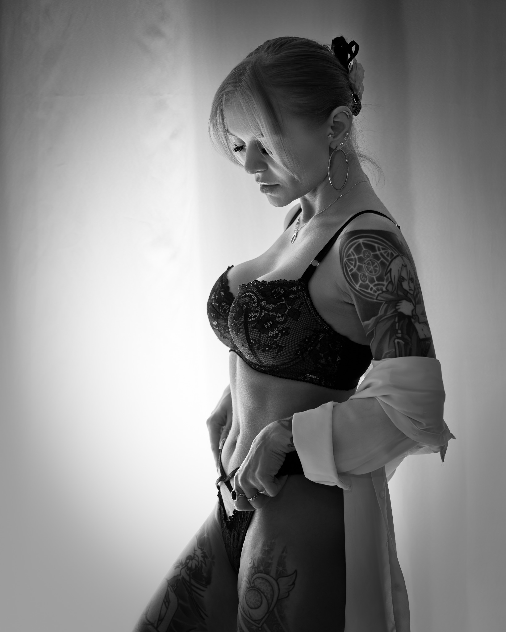 Czarno-białe zdjęcie kobiety w bieliźnie, z tatuażami na ramieniu i udzie, spoglądającej w dół. Ujęcie w półprofilu, z delikatnym oświetleniem i subtelnym tłem.