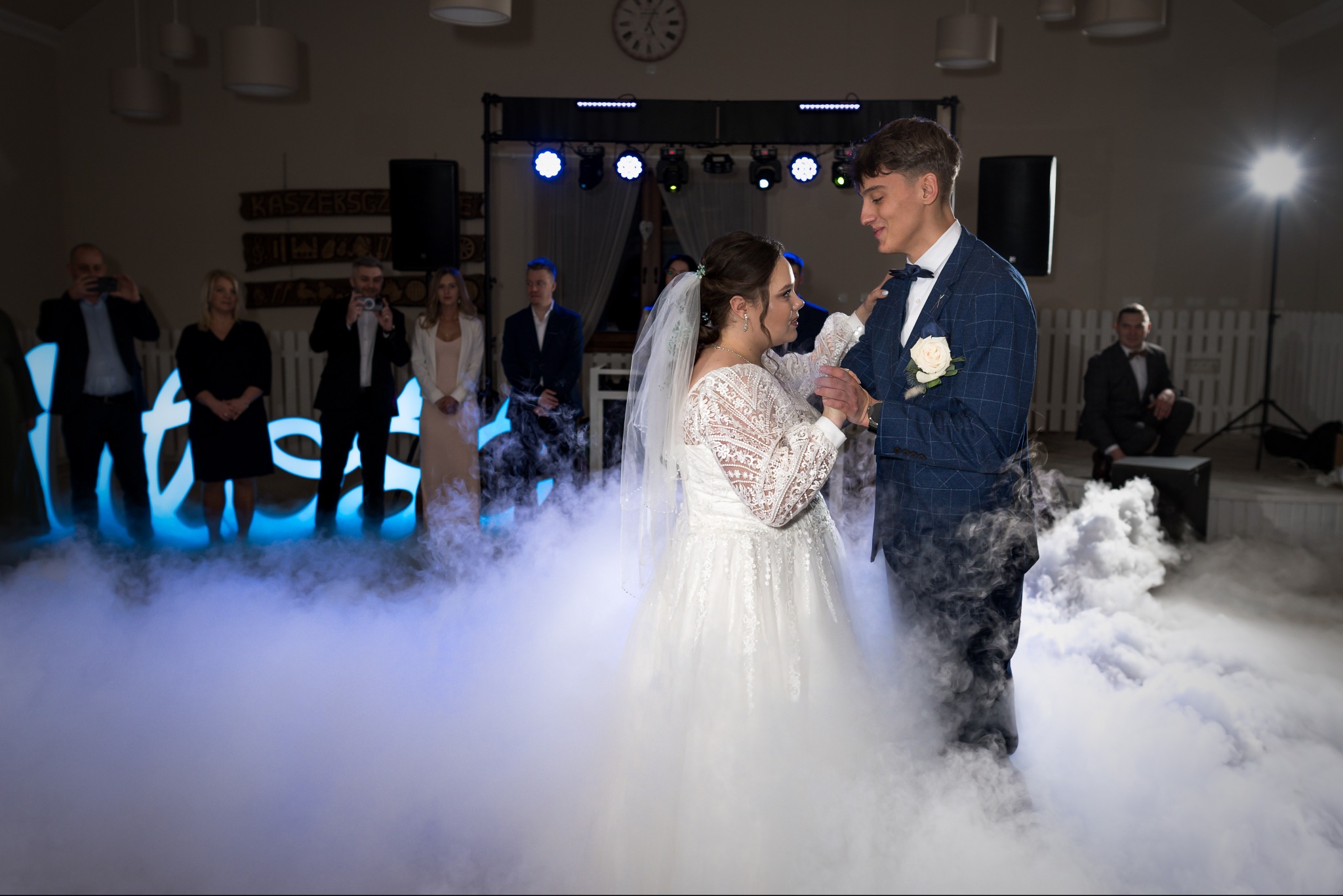 Para młoda w tańcu, otoczona dymem na parkiecie. W tle goście robiący zdjęcia, dekoracje świetlne i zegar na ścianie. Ujęcie z reportażu ślubnego.