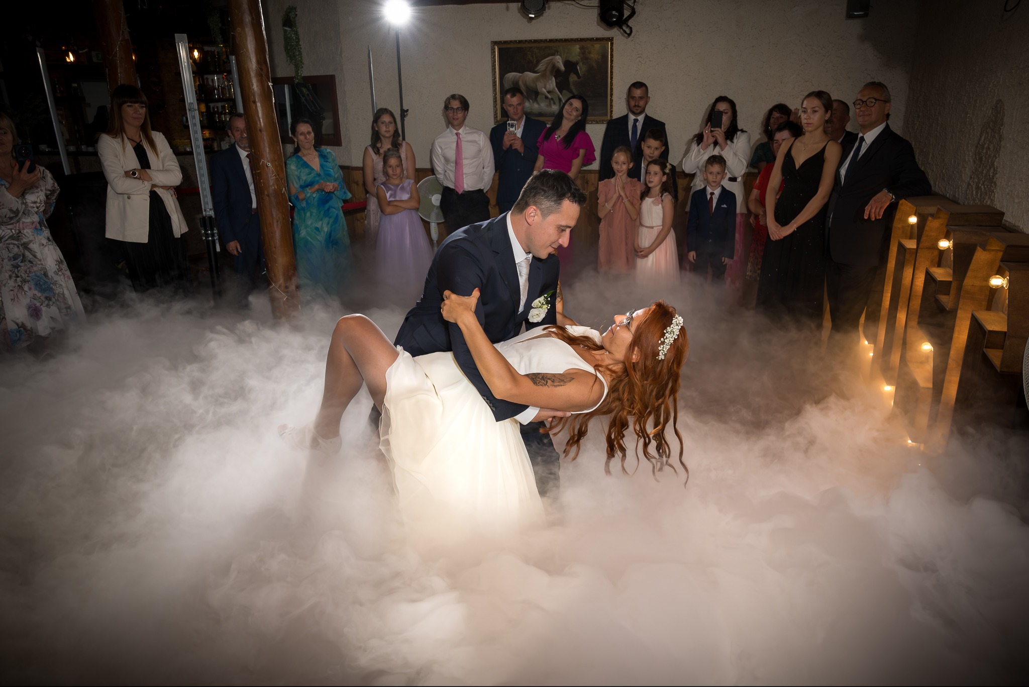 Para młoda w tańcu, unoszona przez pana młodego w dymie na tle gości weselnych. Panna młoda w białej sukni, pan młody w granatowym garniturze. Dekoracja sali.