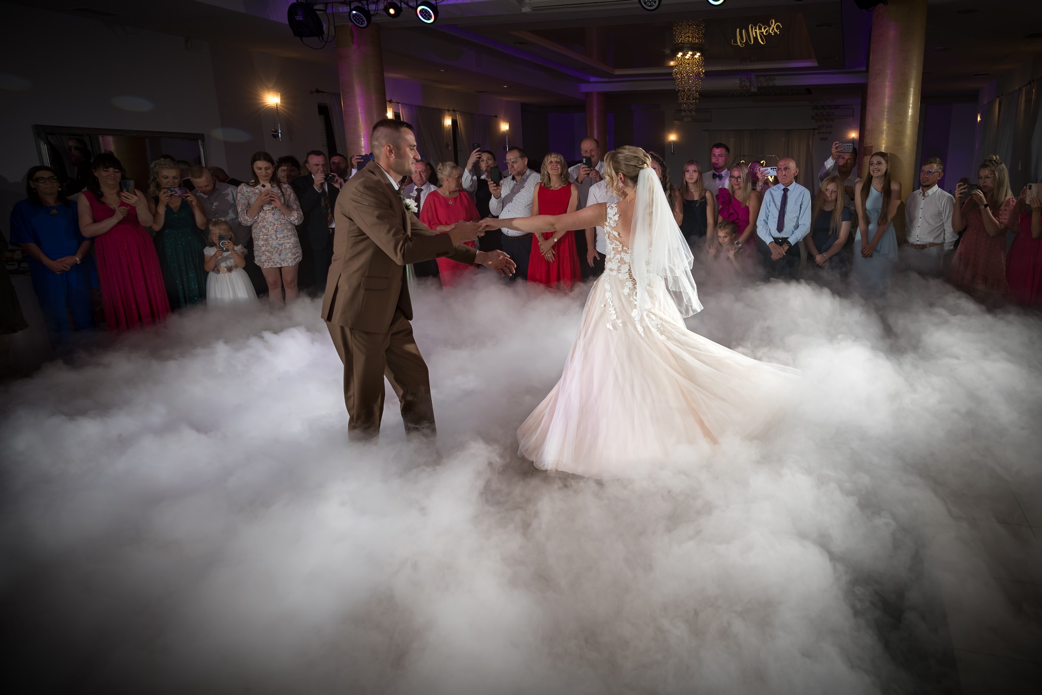 Para młoda w tańcu na parkiecie wypełnionym dymem, otoczona gośćmi weselnymi. Panna młoda w sukni z aplikacjami, pan młody w brązowym garniturze. Dekoracje sali.