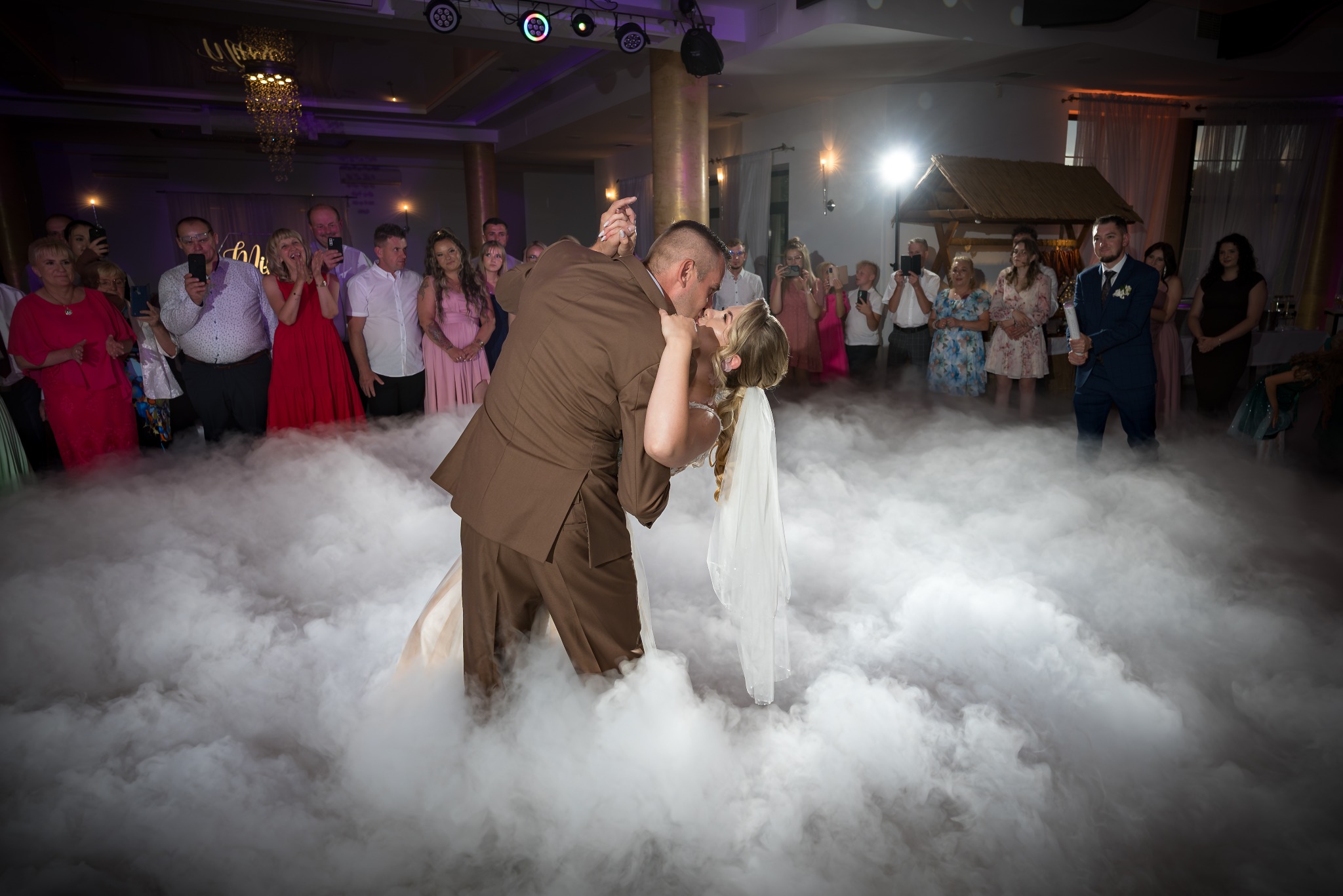Pierwszy taniec pary młodej w objęciach, otulonych sztuczną mgłą, na tle gości weselnych z telefonami i dekoracji sali.