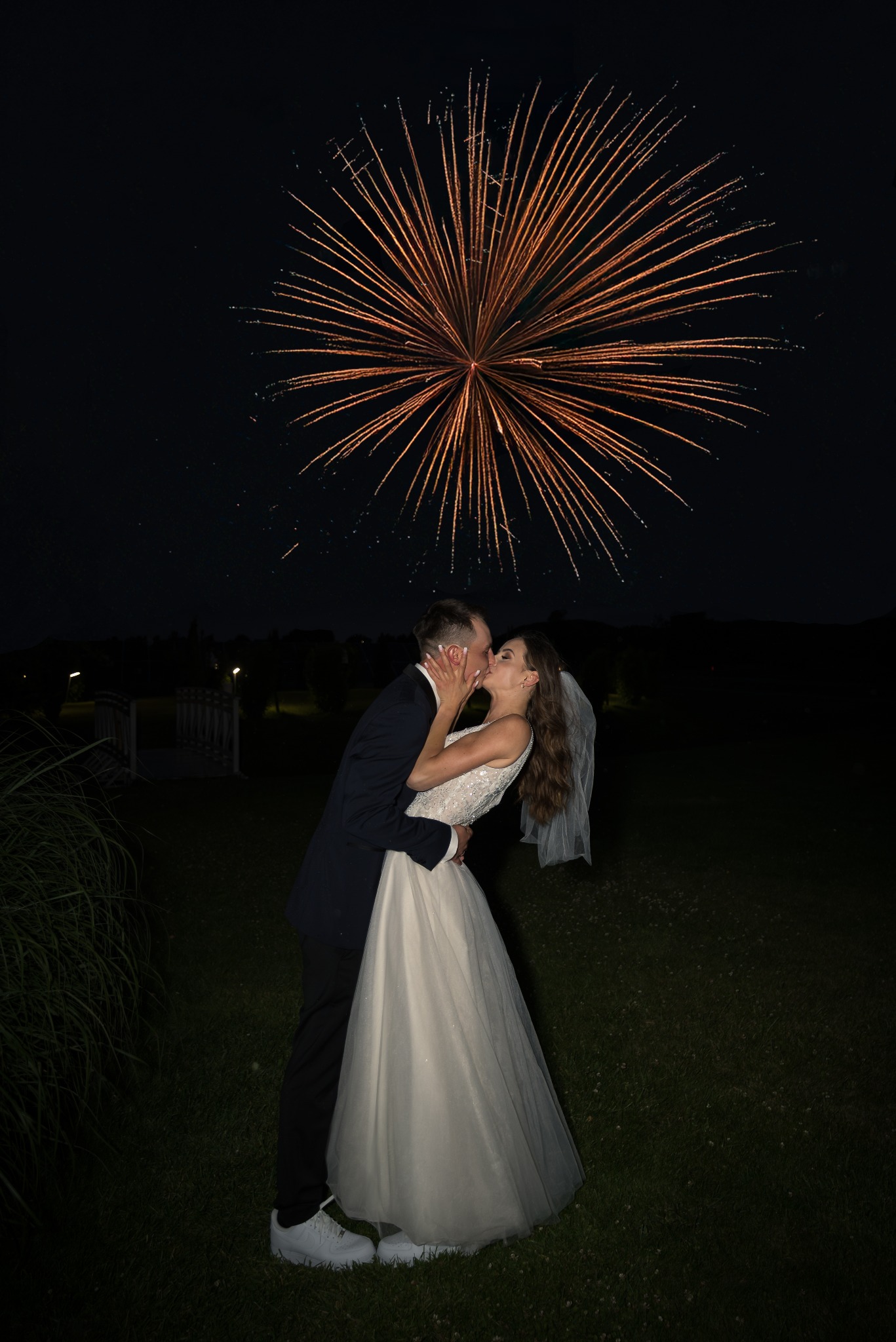 Pocałunek pary młodej na tle nocnego nieba rozświetlonego fajerwerkami. Pan młody w ciemnym garniturze, panna młoda w białej sukni z welonem.