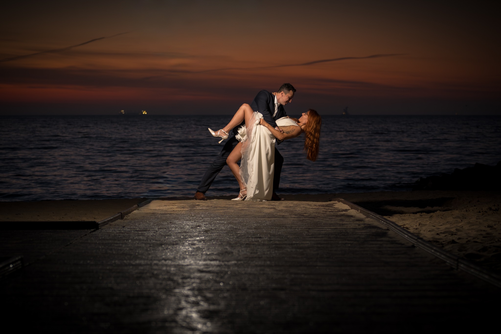 Para młoda w tańcu na tle zachodzącego słońca nad morzem. Pan młody trzyma pannę młodą w ramionach, widoczne światła statków na horyzoncie.