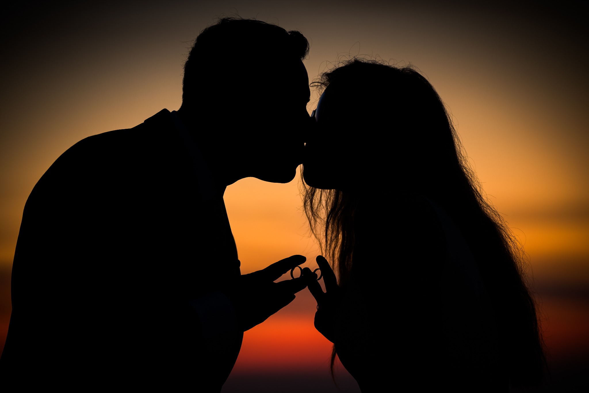 Silueta całującej się pary na tle zachodzącego słońca. W dłoniach trzymają obrączki ślubne, tworząc romantyczny i intymny moment.