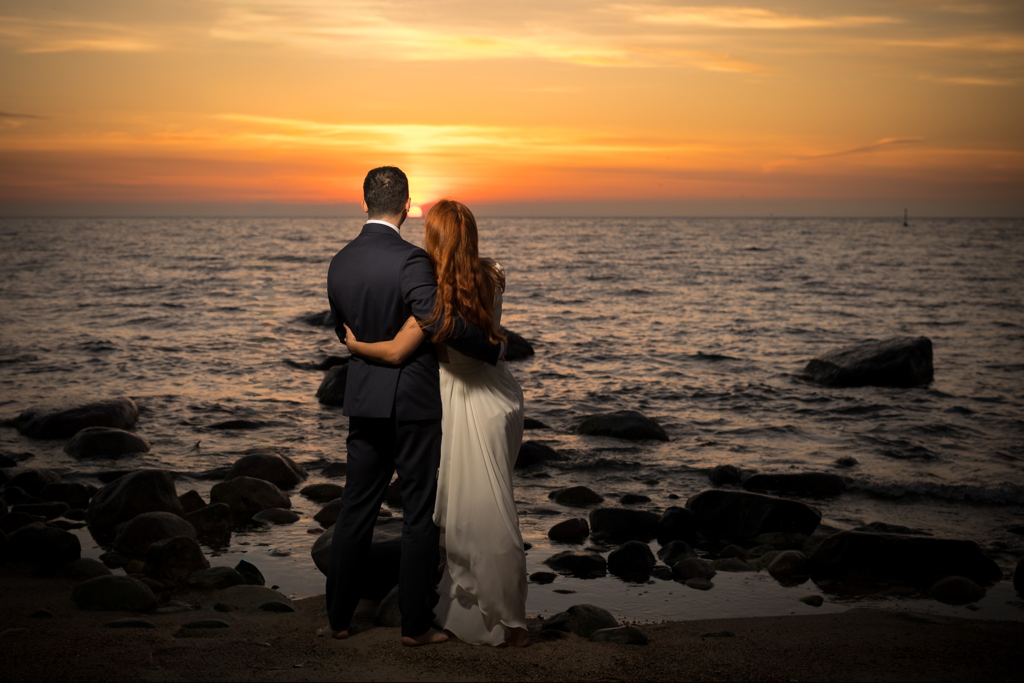 Para młoda obejmuje się na plaży o zachodzie słońca. Pan młody w ciemnym garniturze, panna młoda w białej sukni z rudymi włosami. Romantyczny kadr z widokiem na morze.