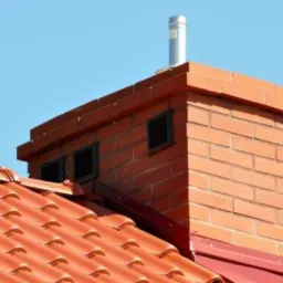 Czerwony dach z ceramicznej dachówki z ceglanym kominem z metalowym wkładem kominowym i kratkami wentylacyjnymi, na tle błękitnego nieba.
