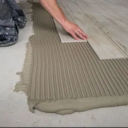 Układanie podłogi z płytek imitujących drewno na świeżo nałożonym kleju z wyraźnymi liniami, widoczne dłonie glazurnika i fragment jego ubrania.