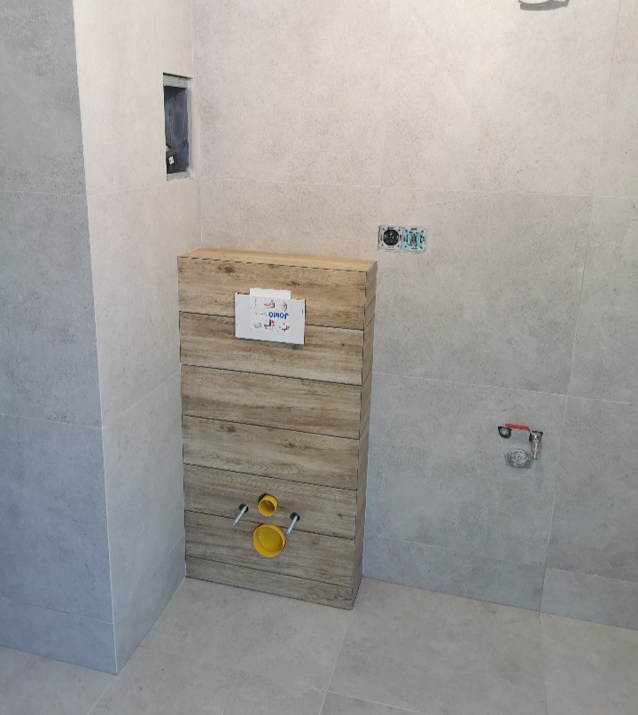 Ściana łazienki wyłożona płytkami imitującymi kamień, z zamontowanym stelażem podtynkowym WC obudowanym płytkami drewnopodobnymi, z widocznymi żółtymi zaślepkami.