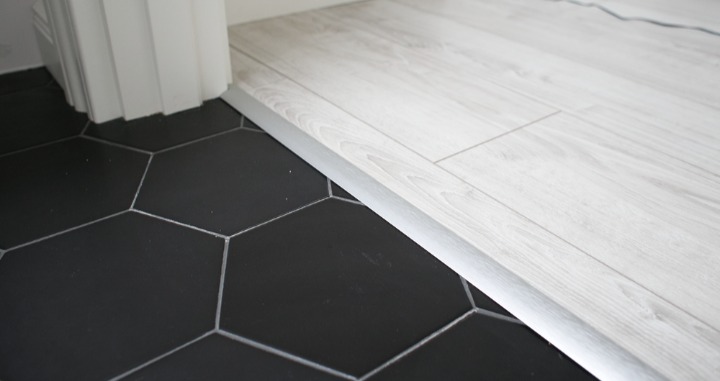 Połączenie podłogi z paneli laminowanych o jasnym odcieniu drewna z czarną, heksagonalną podłogą z płytek, oddzielone srebrną listwą progową.